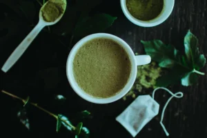 An overhead shot features a frothy cup of green matcha tea centered among loose powder, a tea bag, and fresh leaves. The dark wooden background and soft lighting create a moody, organic atmosphere for the tea preparation.