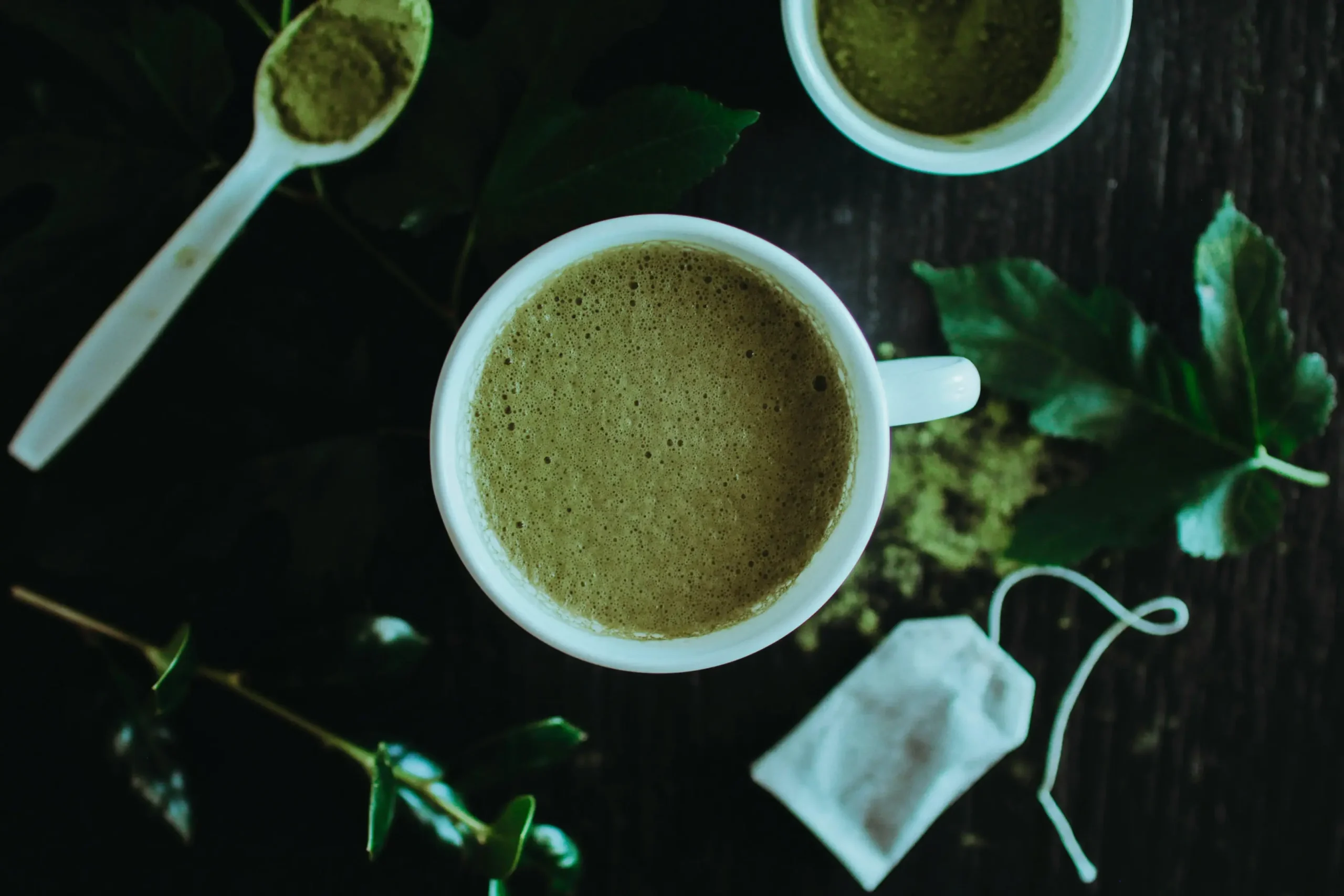 An overhead shot features a frothy cup of green matcha tea centered among loose powder, a tea bag, and fresh leaves. The dark wooden background and soft lighting create a moody, organic atmosphere for the tea preparation.