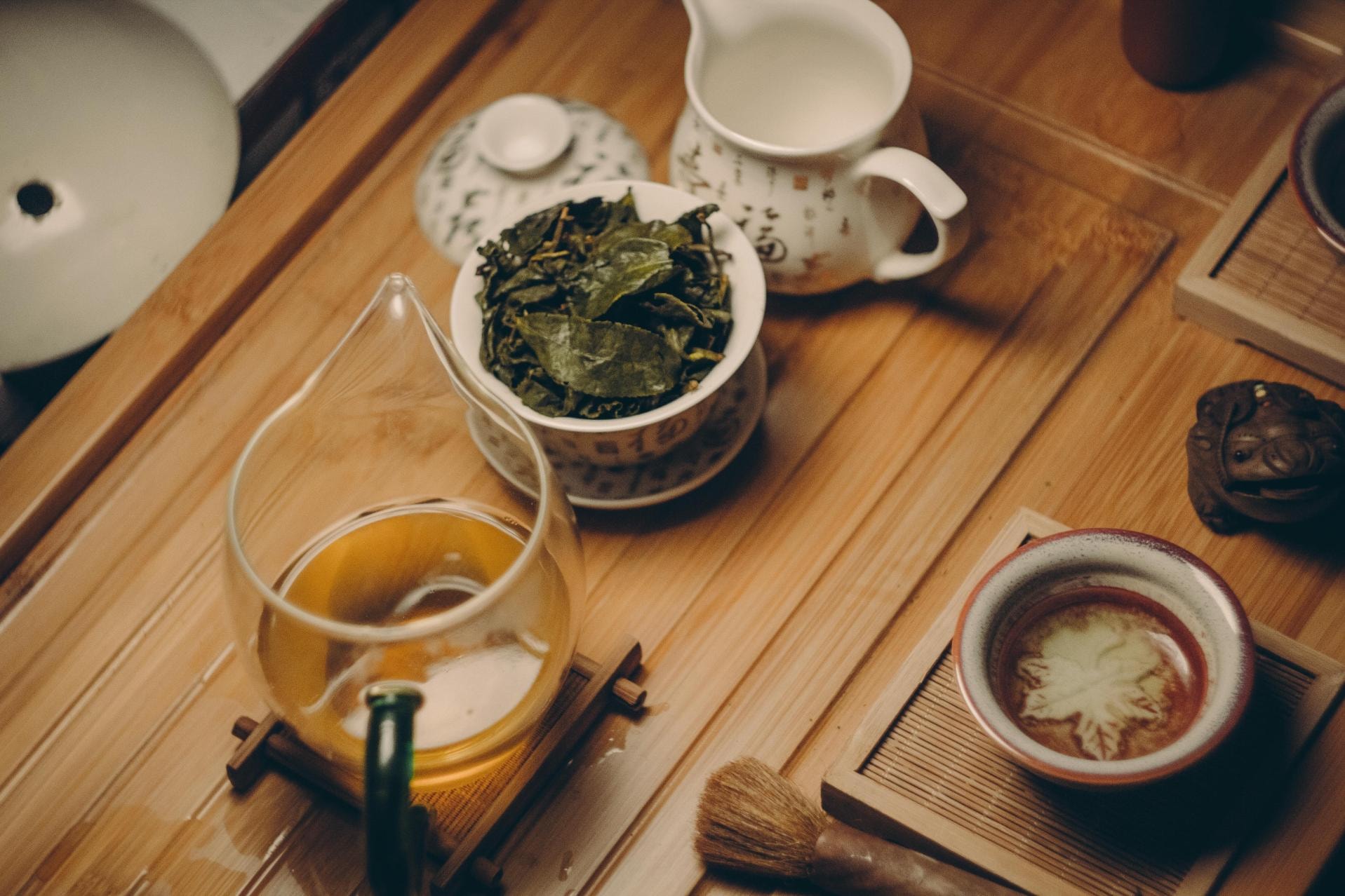 This image displays a traditional Gongfu tea set arranged on a wooden tea tray, featuring a glass pitcher of golden tea and a bowl full of steeped green leaves. Surrounding the main vessels are a small ceramic teacup, a tea pet, and various tools used for a meditative brewing session.