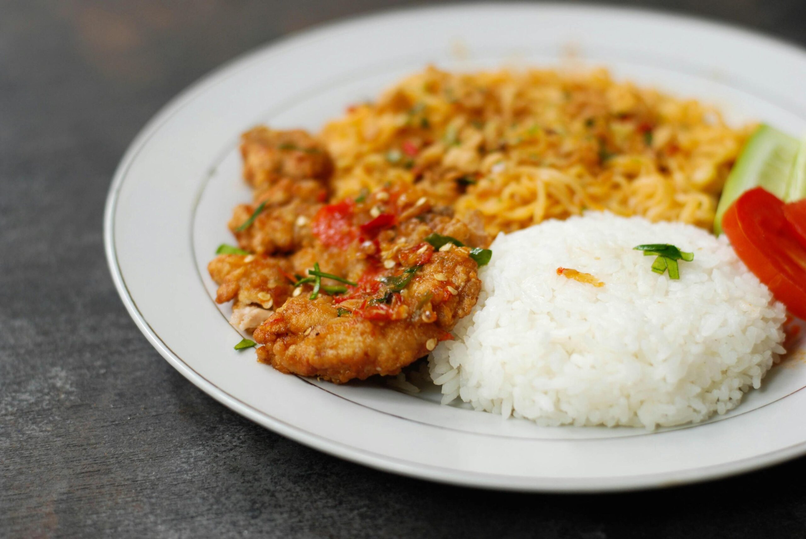 A white plate holds a serving of steamed white rice alongside crispy fried chicken topped with a red chili sambal. The meal is accompanied by stir-fried noodles and fresh garnishes of sliced cucumber and tomato.