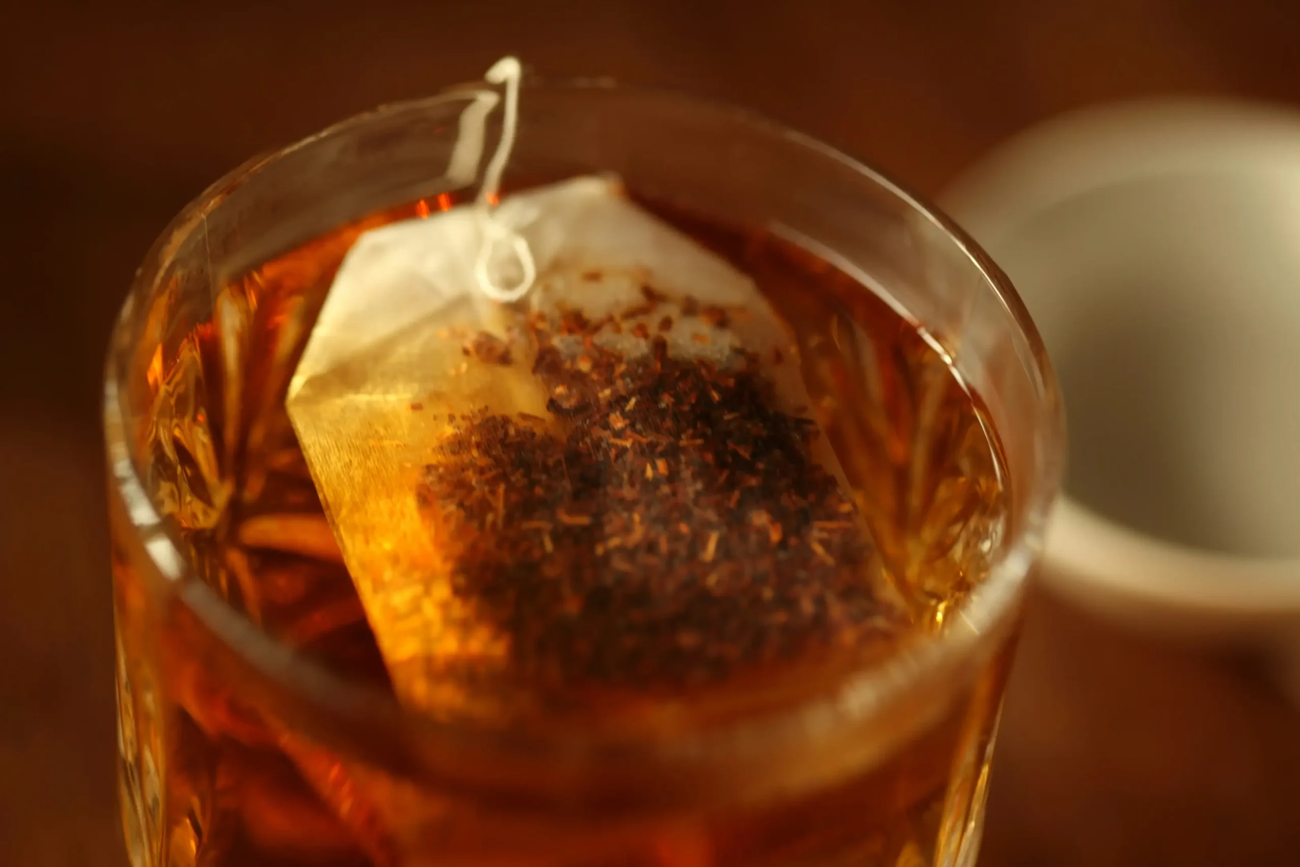 A close-up shot captures a tea bag steeping inside a clear, patterned glass filled with warm, amber-colored liquid. The scene is bathed in a soft, golden glow, creating a cozy and inviting atmosphere.