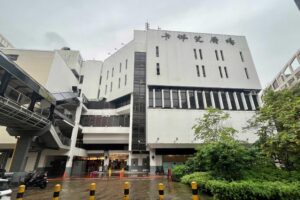 This image shows the exterior of Cuppage Plaza, a multi-story white building in Singapore with its name displayed in both English and Chinese characters. The scene is set on a rainy day, featuring wet pavement, orange traffic cones, and a pedestrian overpass connected to the structure.