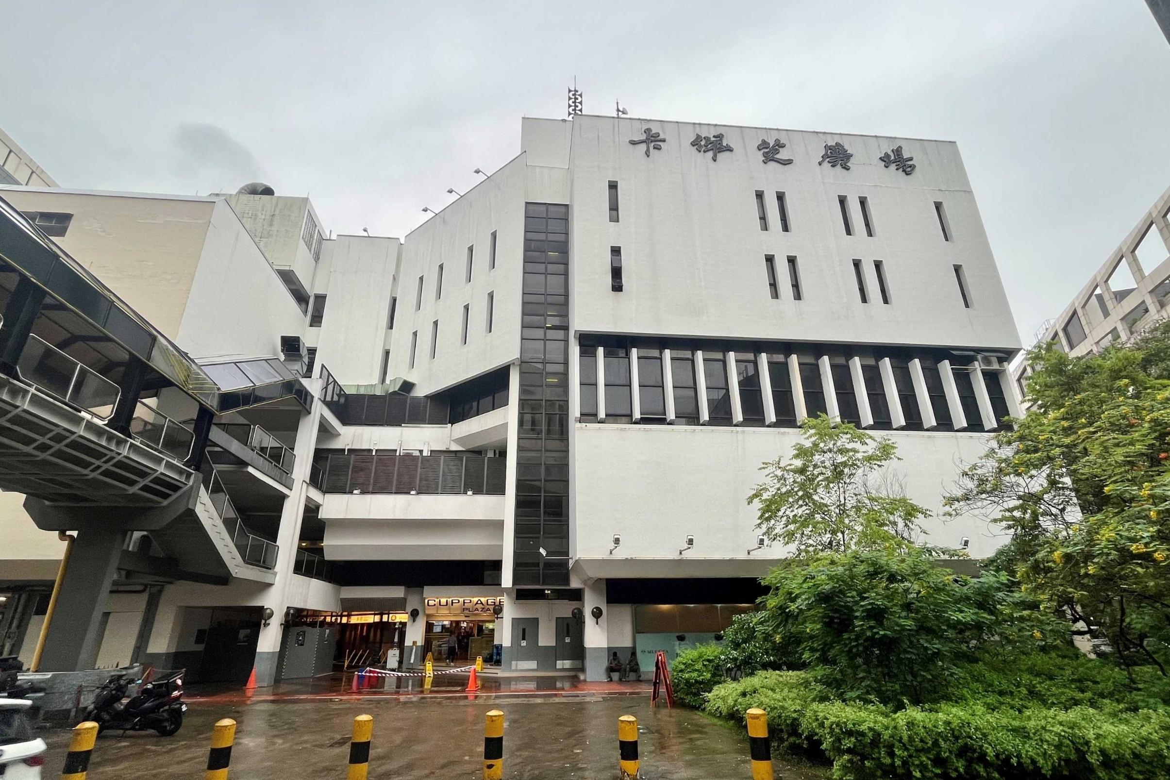 This image shows the exterior of Cuppage Plaza, a multi-story white building in Singapore with its name displayed in both English and Chinese characters. The scene is set on a rainy day, featuring wet pavement, orange traffic cones, and a pedestrian overpass connected to the structure.