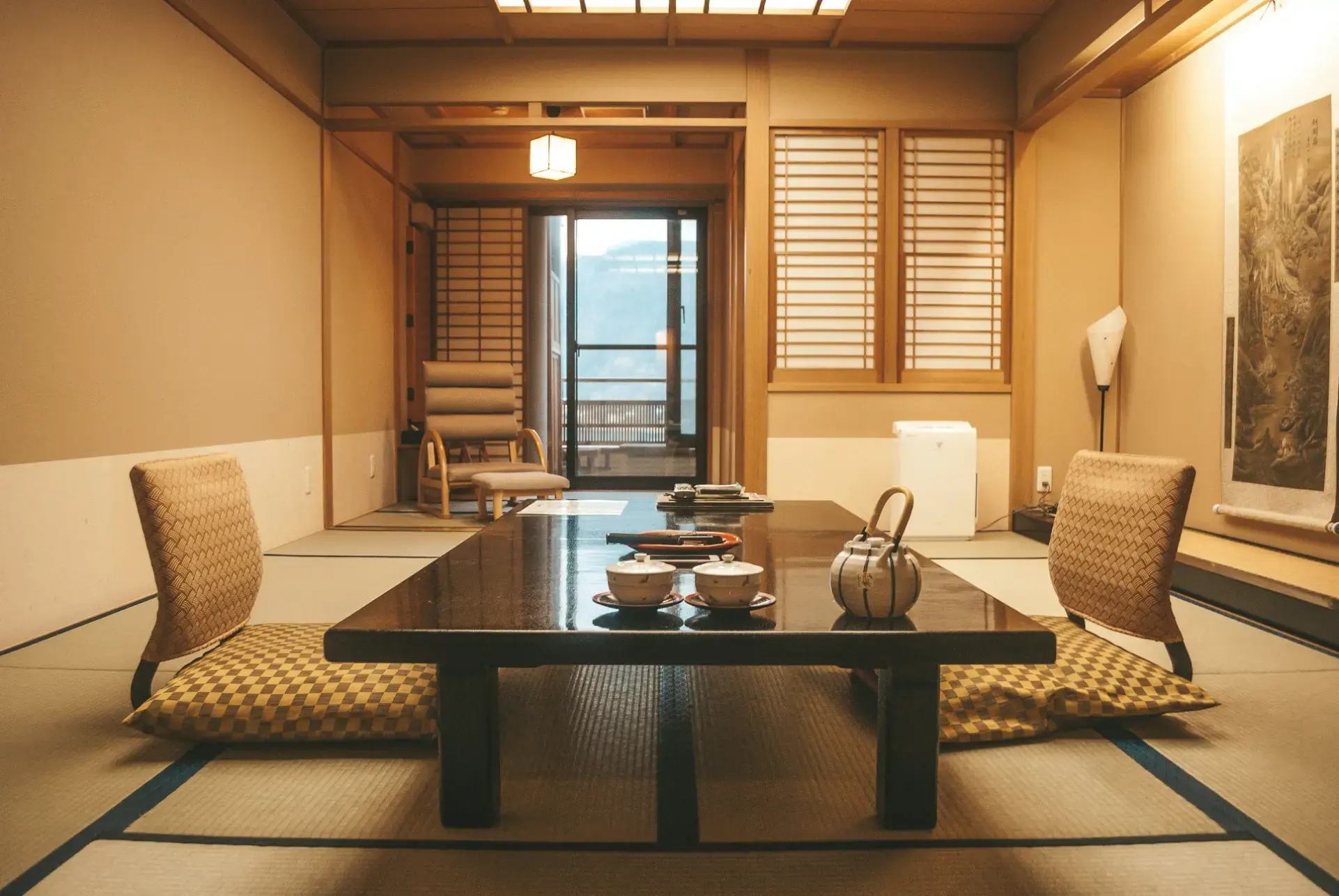 This traditional Japanese room features a low dark wood table set for tea atop tatami mat flooring. Soft, warm lighting illuminates the space, which includes shoji screen doors and floor seating with checkered cushions.