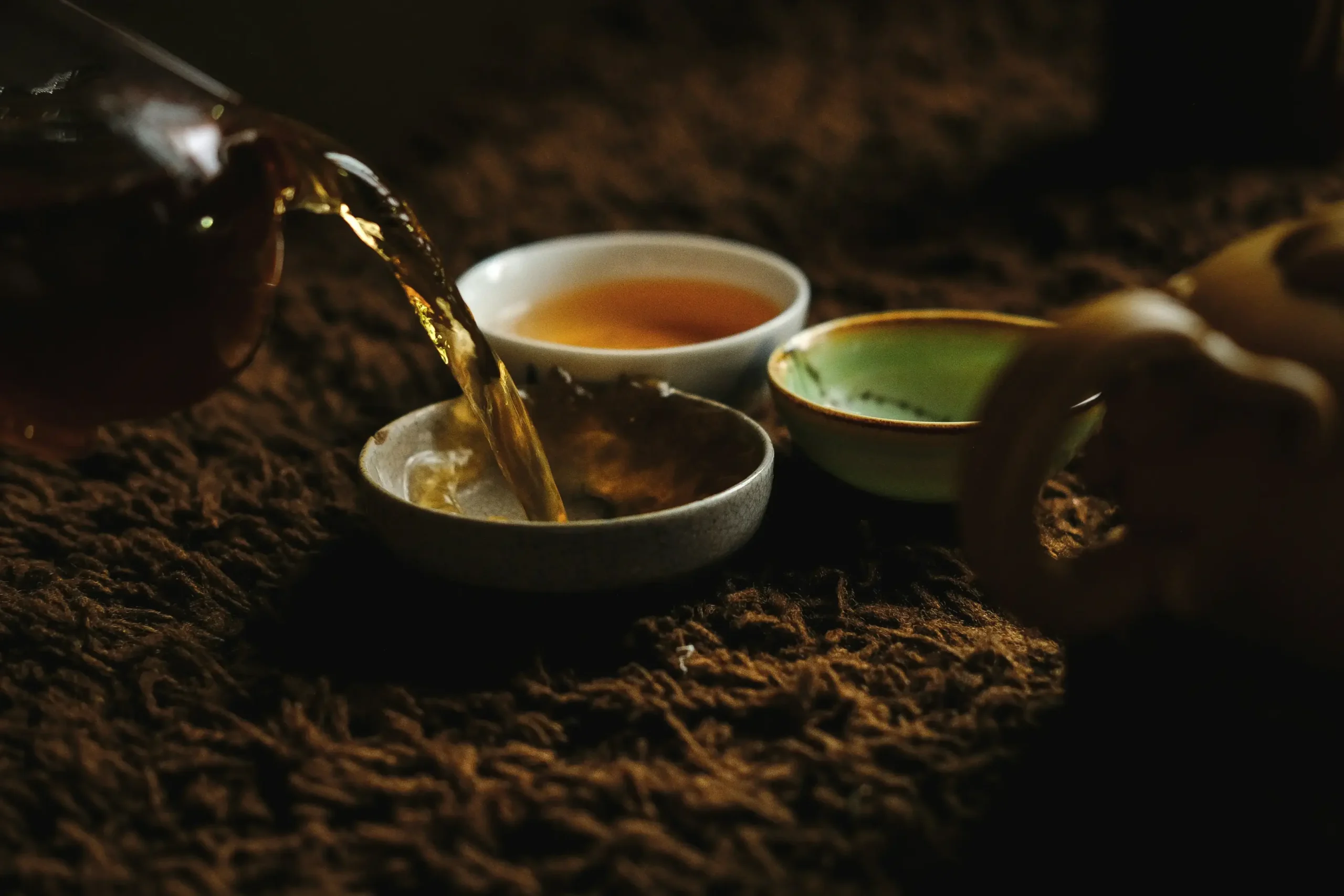 Golden amber tea pours from a glass vessel into a small ceramic bowl, creating a gentle splash. The scene is set in a warm, moody atmosphere with additional tea cups resting on a textured, dark brown surface.