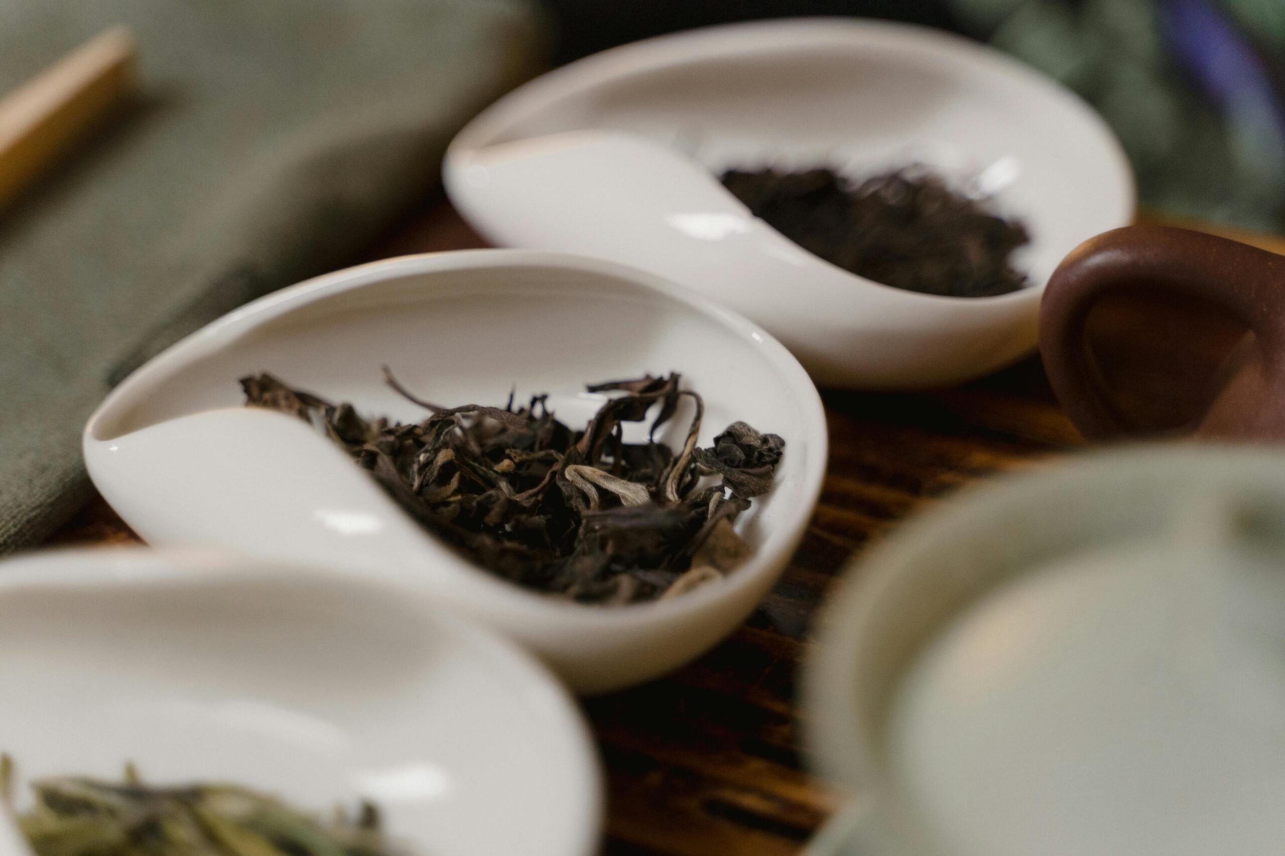 This image captures an array of white, teardrop-shaped ceramic dishes containing various types of loose-leaf tea. The tea leaves are presented on a wooden surface alongside traditional brewing accessories, creating a serene and ritualistic atmosphere.