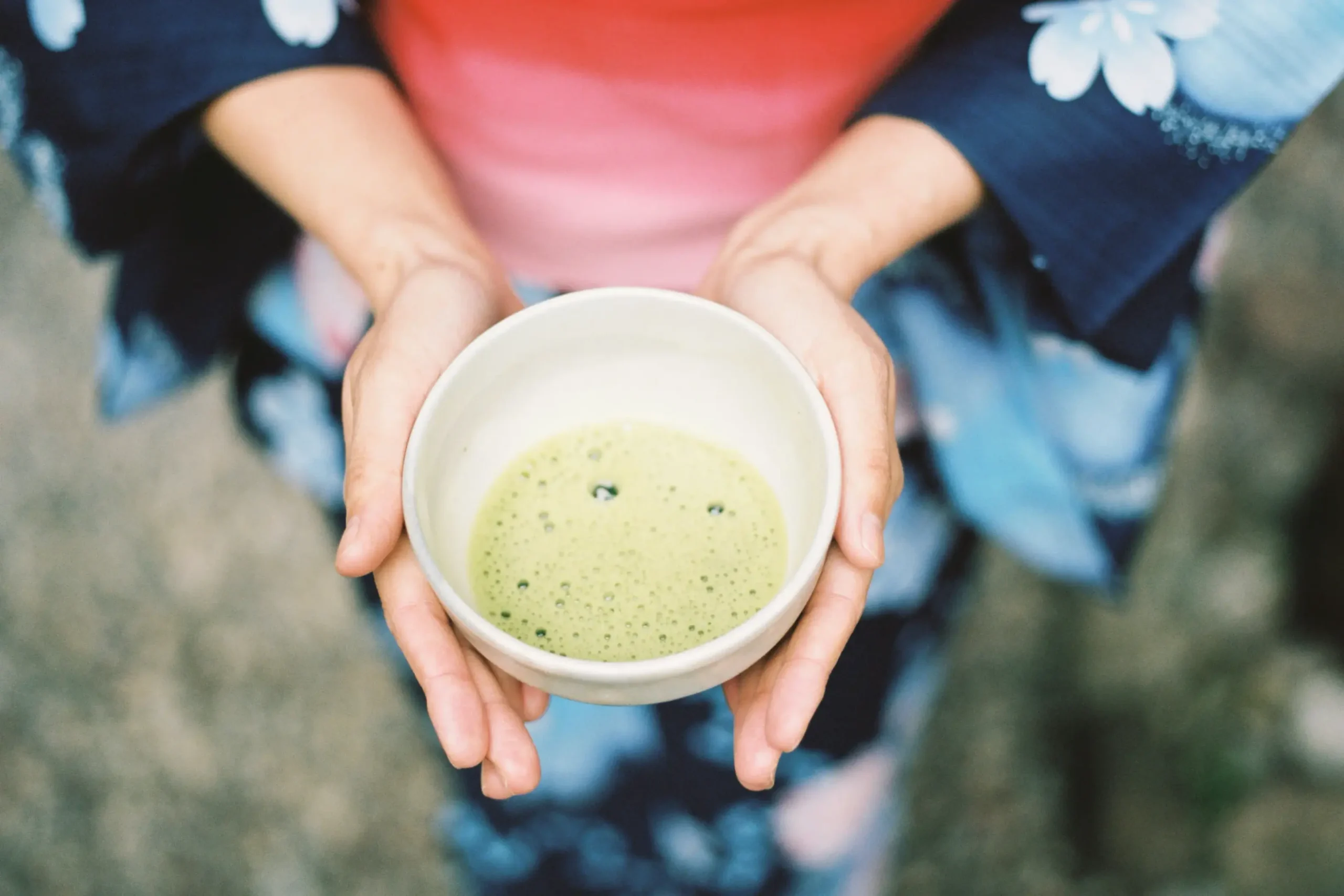 A person dressed in a floral-patterned yukata or kimono holds a ceramic bowl of frothy green matcha with both hands. The overhead perspective captures the serene and traditional essence of a Japanese tea moment.
