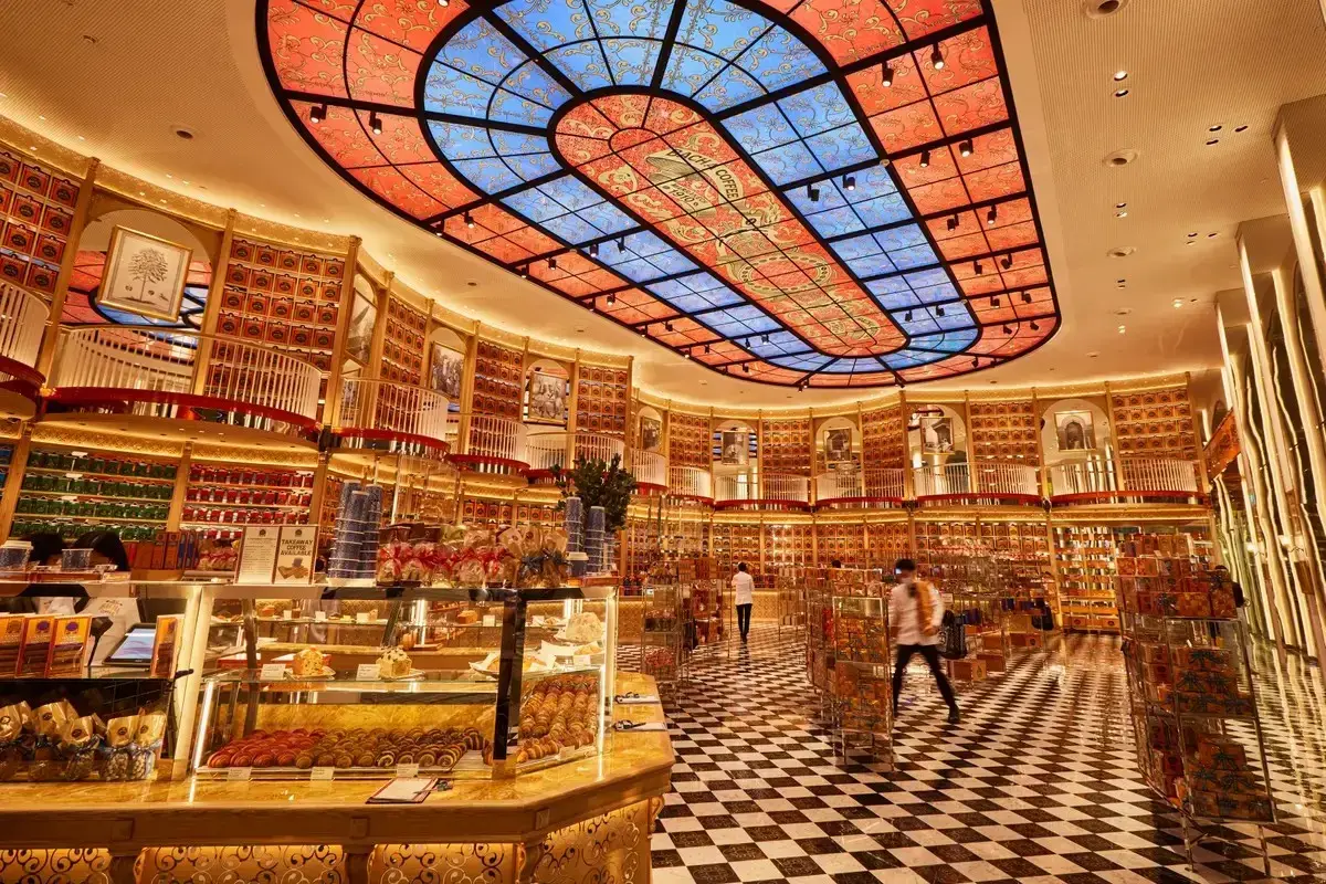 This image showcases the opulent, gold-toned interior of a Bacha Coffee boutique, featuring a striking stained-glass ceiling and a classic black-and-white checkered floor. The walls are lined with floor-to-ceiling shelves of signature orange coffee canisters, while a polished counter displays an array of fresh pastries.