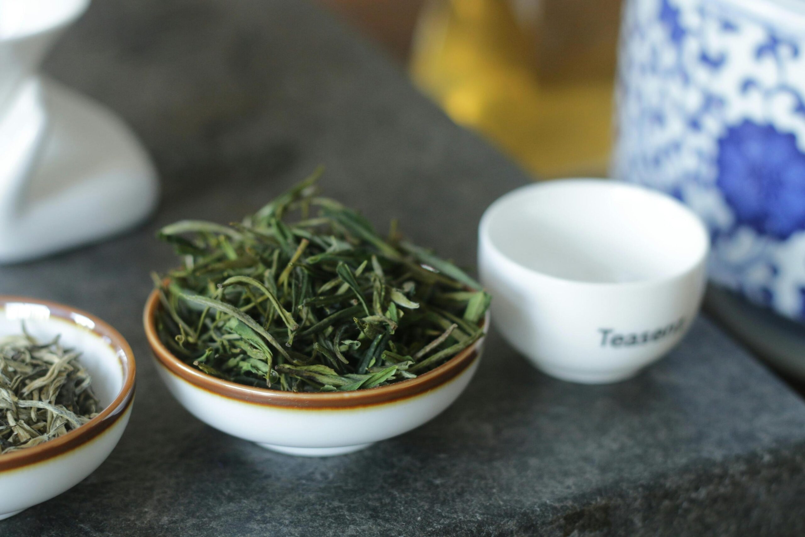 A small ceramic bowl filled with dried green tea leaves sits prominently on a dark stone surface. Beside it, a white tasting cup and a blue-and-white patterned vessel complete the traditional tea preparation scene.