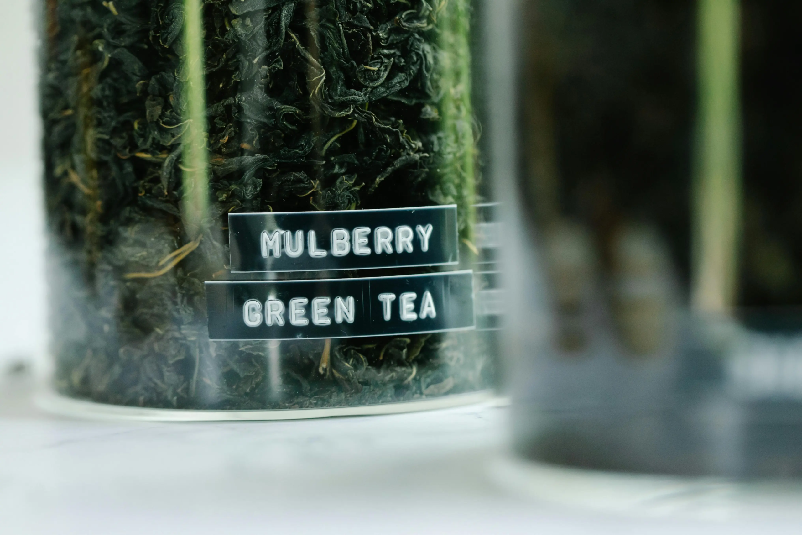 A clear glass jar is filled to the brim with dark, tightly curled dried tea leaves. Two black embossed labels on the front of the container identify the contents as Mulberry Green Tea.