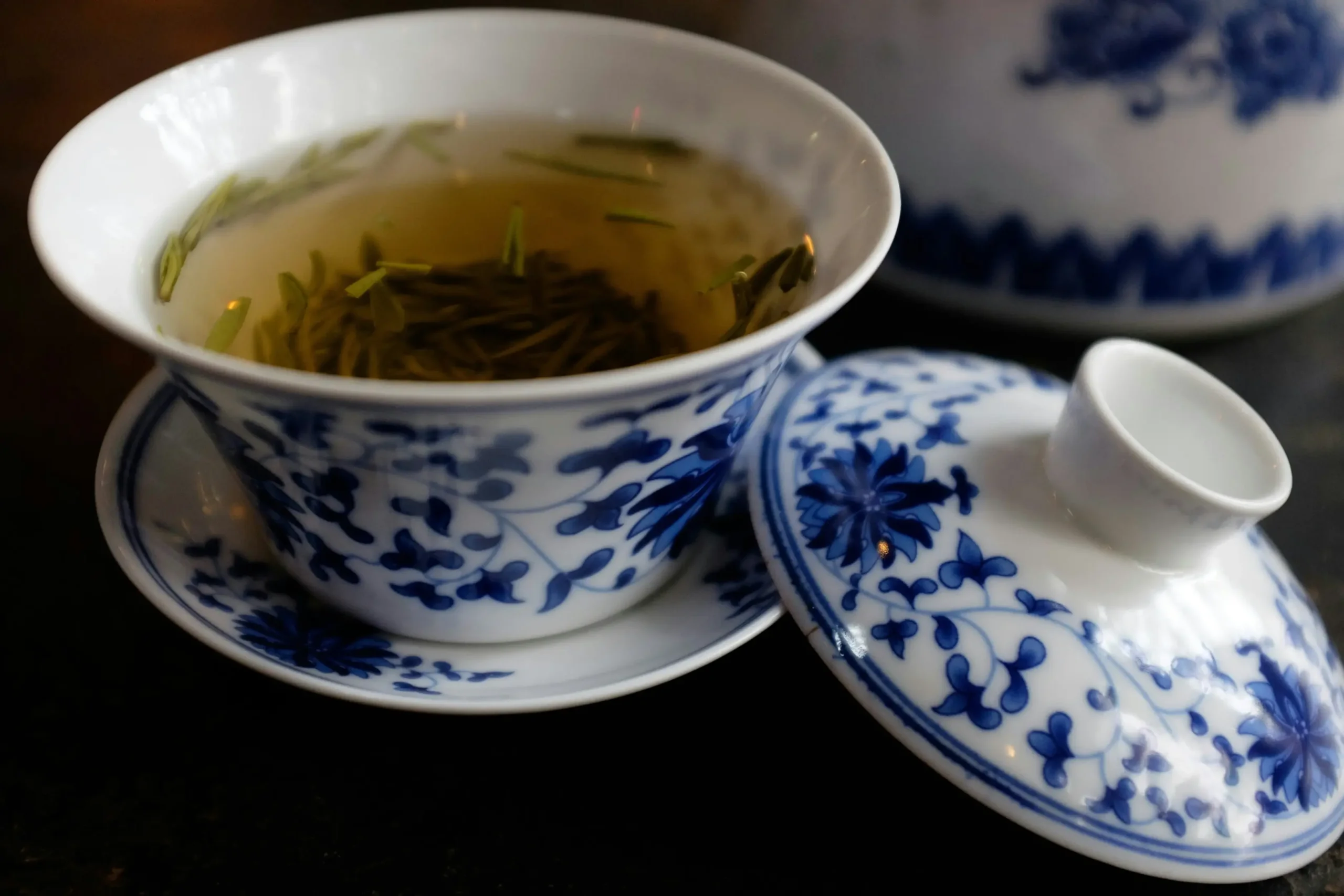 A blue and white floral lidded tea bowl, known as a gaiwan, sits on a matching saucer filled with steeping green tea leaves. The delicate porcelain set features intricate traditional patterns and is positioned next to a similarly decorated teapot in the background.