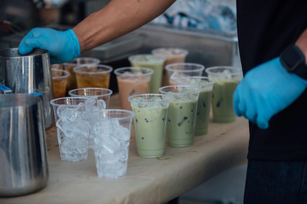 A person wearing blue gloves prepares a large order of iced drinks, including matcha and milk tea, on a paper-covered table. Several clear plastic cups are already filled with ice and colorful beverages, while metal pitchers sit ready for use on the side.