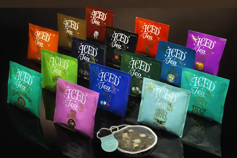 A vibrant collection of TWG Iced Teabags is arranged in neat rows on a reflective black surface, showcasing a variety of colorful packaging and unique flavors. In the foreground, an oversized, round silk teabag is displayed out of its pouch, revealing the detailed blend of tea leaves and dried flowers inside.