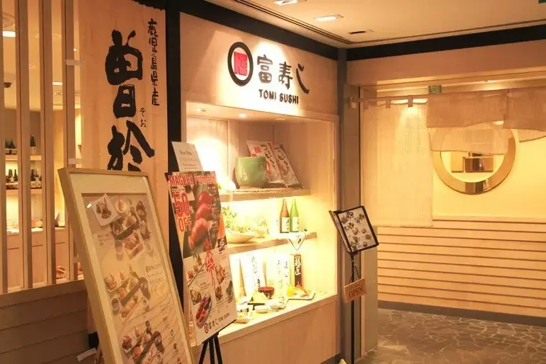 The warm, lit storefront of Tomi Sushi features traditional Japanese design elements, including a light wood exterior, calligraphic signage, and a linen noren curtain at the entrance. A promotional stand and display case showcase various menu items and sake bottles, inviting guests into the clean, modern dining space.