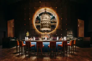 This upscale bar features a curved counter with orange barstools positioned beneath a large circular opening that reveals an ornate, multi-faced bronze bell. The dark, atmospheric space is accented by warm lighting and a textured wall, creating a sophisticated and mystical ambiance.