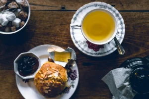 A top-down view shows a rustic wooden table set with a floral teacup of golden tea, a teapot, and a bowl of sugar cubes. Beside the tea sits a plate featuring a sliced scone topped with slivered almonds, accompanied by small servings of jam and butter.