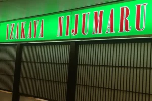 This image shows a brightly lit, neon green sign for a Japanese restaurant called "Izakaya Nijumaru," featuring bold red lettering with white outlines. Below the sign, traditional-style dark wooden lattice screens with frosted glass panels form a storefront or partition.