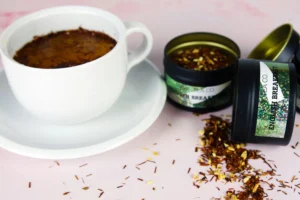 A white ceramic teacup filled with dark amber tea sits on a matching saucer alongside several black tins of loose-leaf English Breakfast tea. Loose tea leaves are scattered across the light pink surface, creating a textured and aromatic scene.