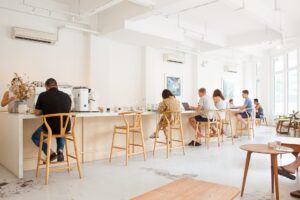 This image features a bright, minimalist café interior with white walls, high ceilings, and patrons seated along a long light-wood counter. Natural light floods the space from large windows, highlighting the clean aesthetic of the wooden wishbone chairs and simple furniture.