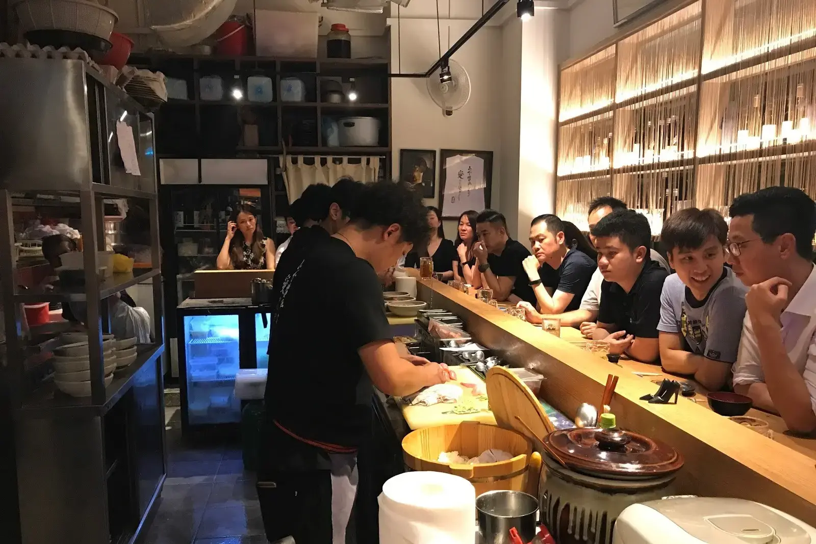 A chef meticulously prepares dishes behind a wooden counter in a lively, narrow Japanese-style eatery. Patrons sit closely together along the bar, watching the preparation in a warm, dimly lit atmosphere filled with kitchenware and authentic decor.