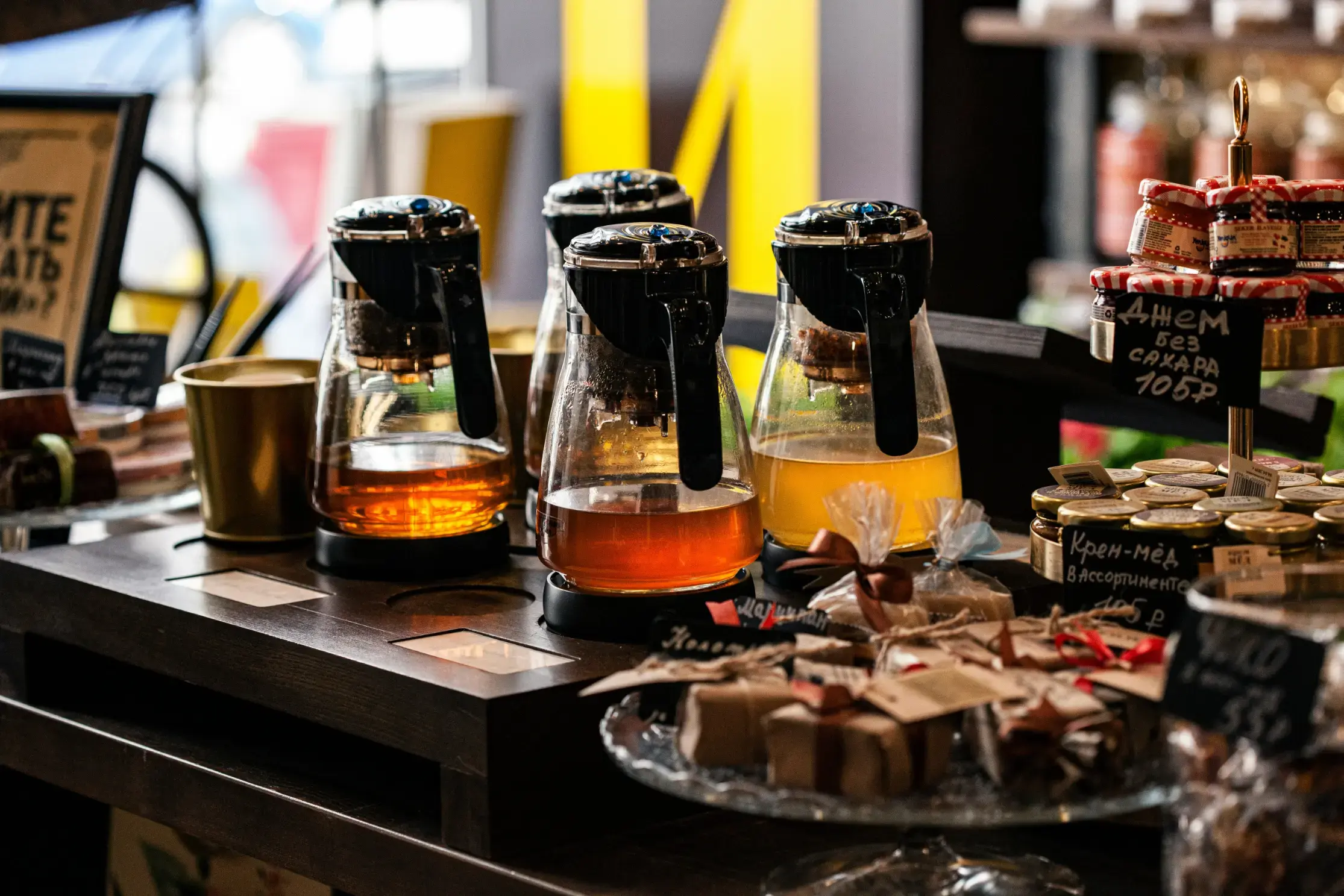 Several glass teapots filled with various golden and amber-hued teas sit on a wooden display counter alongside small jars of honey and jam. The warm, inviting scene is captured with a shallow depth of field, highlighting the artisanal gift sets and tea brewing equipment in a cozy shop setting. Would you like me to help you write a marketing caption for this photo?