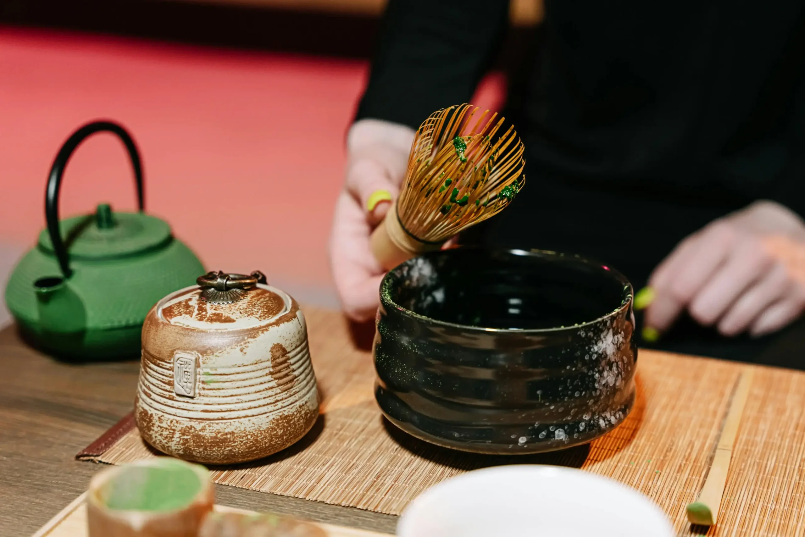 A person prepares ceremonial matcha, lifting a bamboo whisk stained with vibrant green tea from a dark, textured ceramic bowl. The scene is set with traditional tea tools, including a rustic clay canister and a green cast-iron teapot resting on a bamboo mat.
