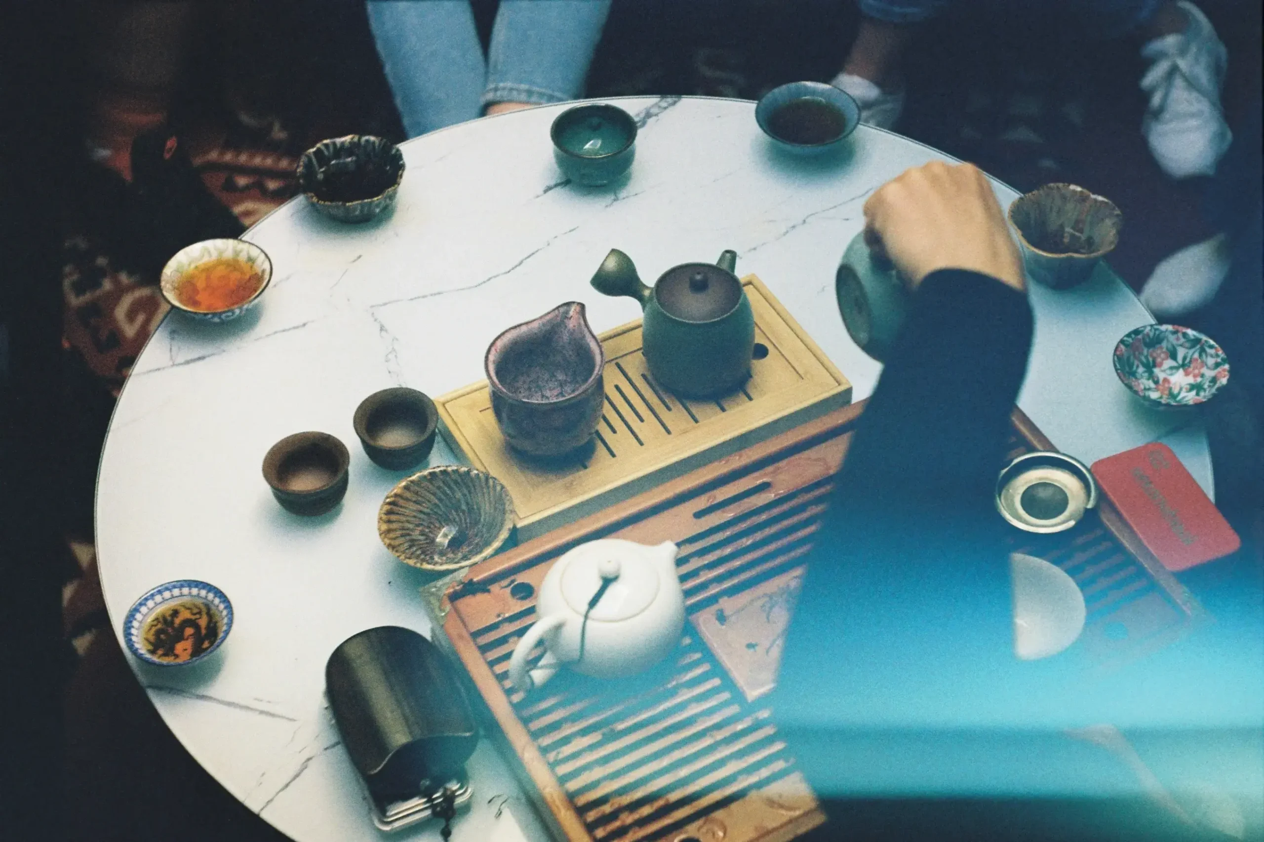 A person pours tea from a ceramic pot into an array of colorful cups arranged on a white marble table. The overhead shot captures a cozy gathering, featuring wooden tea trays and vintage-style film grain that adds a nostalgic warmth to the scene.