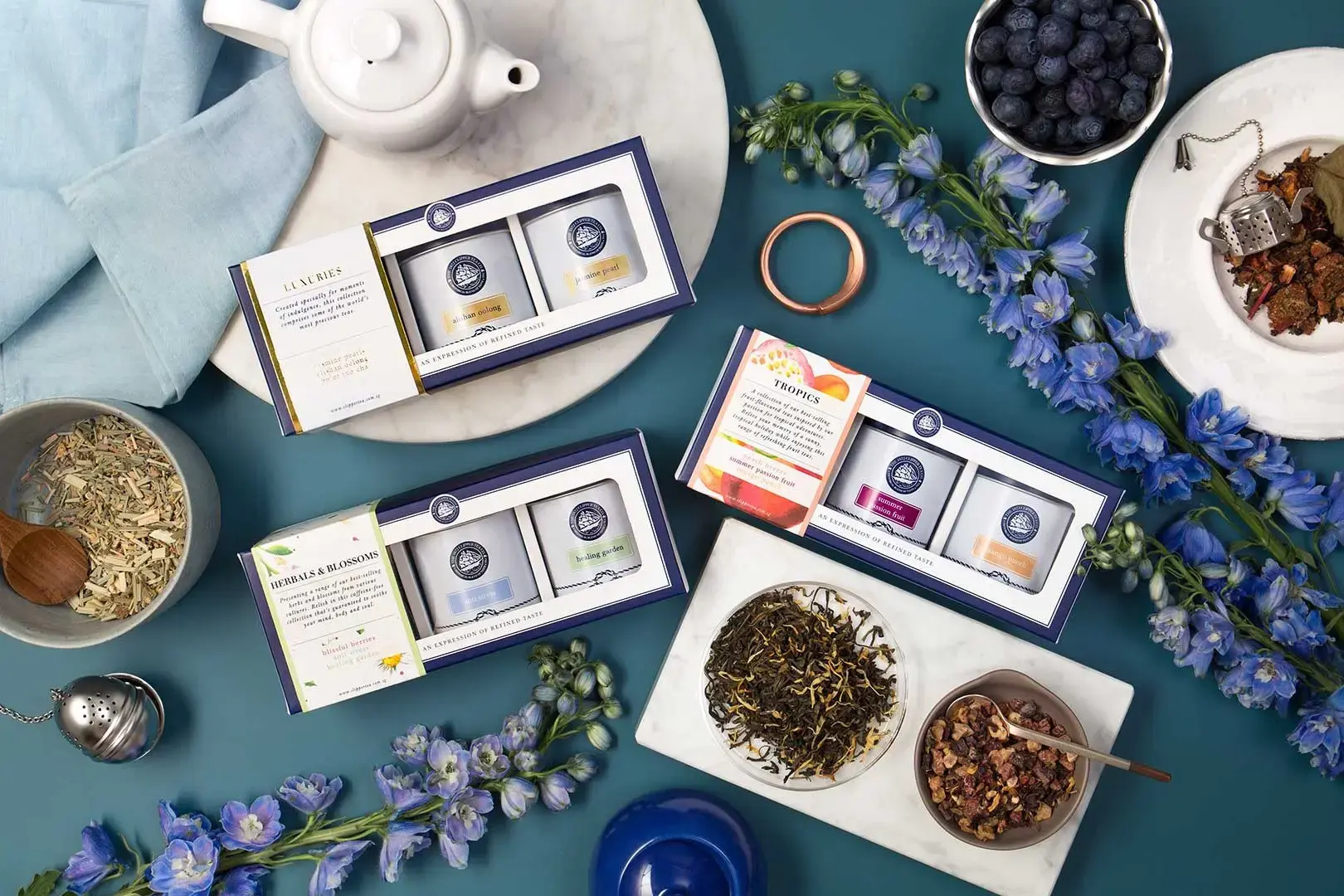 This flat-lay composition showcases three tea gift sets from The 1872 Clipper Tea Co., arranged elegantly alongside loose leaves, fresh blueberries, and blue delphiniums. The scene is set on a deep teal surface, complemented by white marble accents and a ceramic teapot to evoke a sophisticated brewing experience.