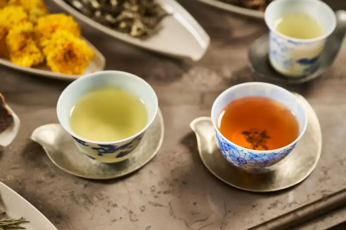 A curated selection of traditional tea is presented in delicate blue-and-white porcelain cups resting on elegant metallic saucers. The warm, overhead lighting highlights the rich colors of the brewed tea alongside bowls of loose tea leaves and vibrant yellow blossoms.
