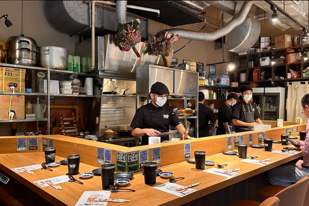 An indoor shot of a bustling Japanese restaurant kitchen and counter seating area. In the foreground, an L-shaped wooden counter is meticulously set for diners with black tumblers, small blue-and-white soy sauce dishes, chopsticks, and menus, while small square QR code signs are pasted along the counter's inner rim. Behind the counter, three kitchen staff members wearing black uniforms, caps, and white face masks are actively preparing food. The background reveals an industrial-style kitchen filled with stainless steel shelves, large cooking pots, stacked containers, exposed overhead ventilation ducts, and a wooden shelf stocked with various kitchen supplies, creating an authentic, high-energy dining atmosphere.
