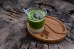 An iced matcha latte with distinct green and white layers is served in a clear glass on a rustic wooden coaster. The drink features a large sphere of ice and sits atop a weathered, textured tree stump.