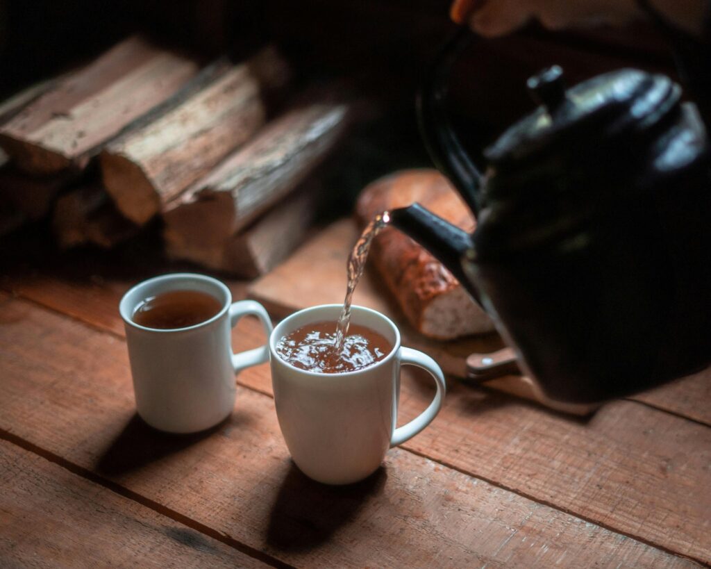 A black kettle pours a warm, amber-colored liquid into one of two white mugs resting on a rustic wooden table. The cozy, dimly lit scene is set against a background of stacked firewood and a loaf of bread.
