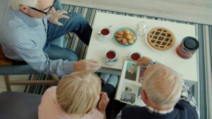 An elderly couple sits together at a coffee table, sharing tea and pastries while looking through a vintage photo album. The overhead shot captures a nostalgic moment as they lean in to view old photographs alongside a younger man.