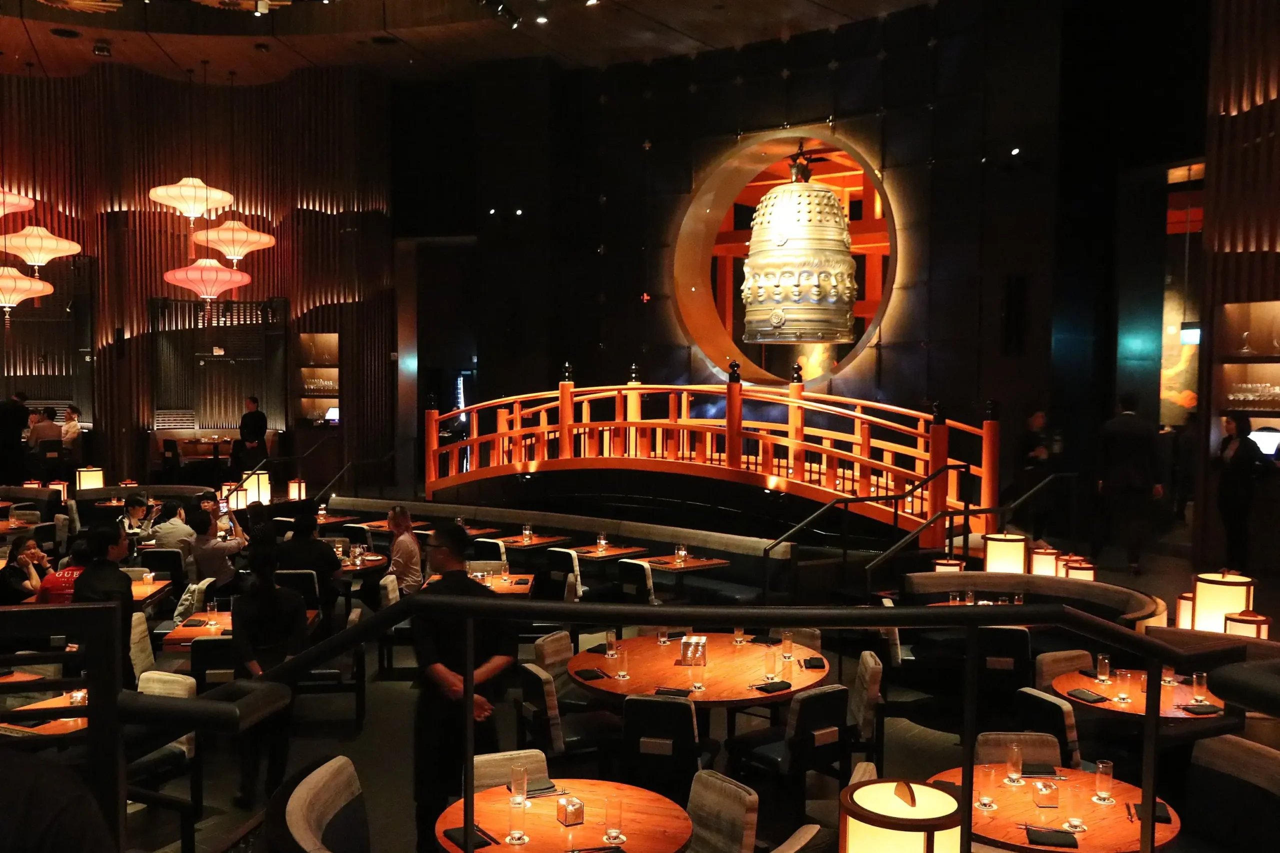 The interior of this high-end restaurant features a striking red bridge that spans across the dining area toward a massive, ornate golden bell suspended in a circular alcove. Warm, atmospheric lighting from paper lanterns and glow-lit tables complements the dark, sophisticated architectural details of the multi-level space.