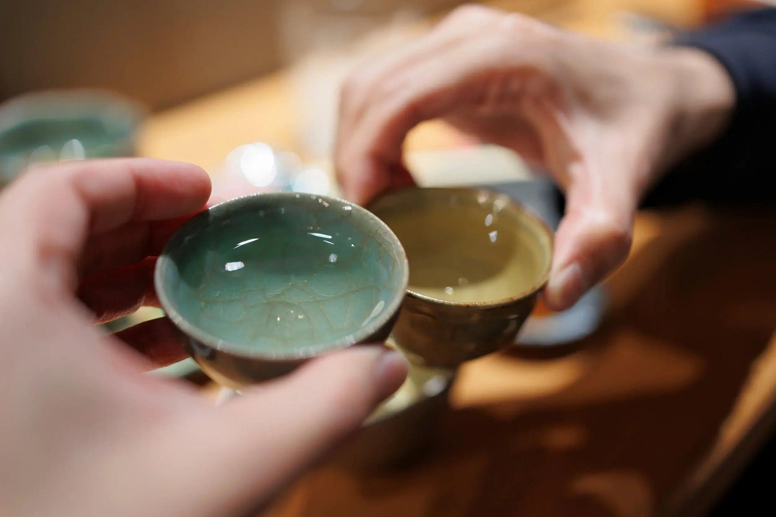 Two hands hold up small, ceramic tea cups in a toast, showcasing the delicate crazing pattern in the pale green glaze of the foreground cup. The shallow depth of field creates a soft, warm bokeh background that emphasizes the intimate moment of sharing a drink.