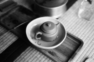 A small, clay teapot sits centered in a shallow bowl atop a wooden tray and textured mat. The photograph is captured in high-contrast black and white with a grainy, film-like quality.