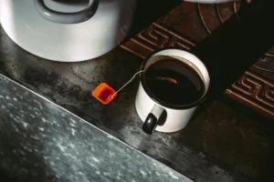 A high-angle shot shows a white enamel mug filled with dark tea, its orange teabag string draped over the side onto a rustic, weathered surface. Next to the mug sits a large white pot, all illuminated by a dramatic, sharp shadow slicing across the scene.