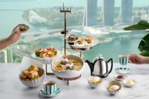 An elegant high tea spread features an assortment of sweet and savory pastries displayed on tiered stands next to a silver teapot and striped teacups. The table is set against a floor-to-ceiling window offering a stunning, elevated view of Singapore's Marina Bay waterfront.