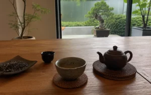 A serene, eye-level shot captures a traditional tea setting arranged on a smooth, warm-toned wooden table, evoking a minimalist and peaceful atmosphere. In the foreground, a dark clay Yixing-style teapot rests on a circular woven coaster to the right, balanced by a speckled, light-grey ceramic teacup filled with pale tea sitting on its own matching coaster in the center. To the left, a small, dark teacup stands beside a curved, leaf-shaped dark dish holding a neat pile of loose, dried tea leaves. In the softly blurred background, a large glass window opens up to a tranquil outdoor courtyard, featuring a meticulously manicured bonsai tree in a rectangular planter and lush green shrubbery beneath an overcast sky, framed elegantly by a slender indoor potted tree on the left and a vibrant green plant on the right.