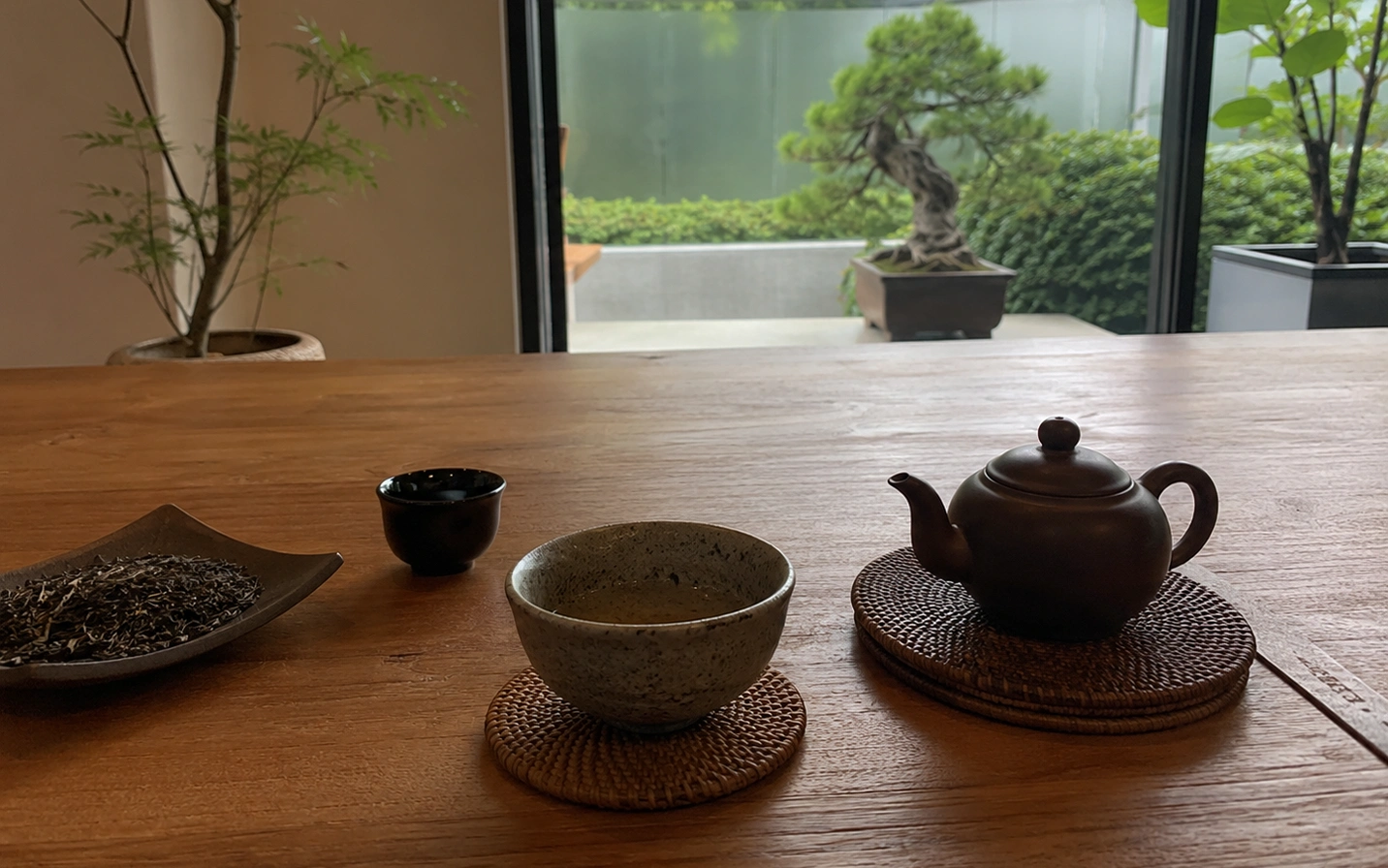 1 - Not Every Expensive Tea Is Actually Better A serene, eye-level shot captures a traditional tea setting arranged on a smooth, warm-toned wooden table, evoking a minimalist and peaceful atmosphere. In the foreground, a dark clay Yixing-style teapot rests on a circular woven coaster to the right, balanced by a speckled, light-grey ceramic teacup filled with pale tea sitting on its own matching coaster in the center. To the left, a small, dark teacup stands beside a curved, leaf-shaped dark dish holding a neat pile of loose, dried tea leaves. In the softly blurred background, a large glass window opens up to a tranquil outdoor courtyard, featuring a meticulously manicured bonsai tree in a rectangular planter and lush green shrubbery beneath an overcast sky, framed elegantly by a slender indoor potted tree on the left and a vibrant green plant on the right.
