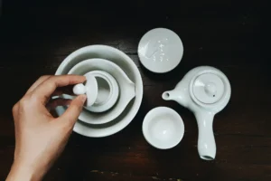 A person’s hand rests on the lid of a small white ceramic teapot nested within several matching bowls. The minimalist tea set is arranged on a dark wooden surface alongside a side-handled teapot and a small cup.