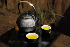 A traditional Japanese cast-iron teapot with a textured, studded surface and a rope-wrapped handle rests on a sleek, black reflective surface, accompanied by two small, dark ceramic cups filled with vibrant green tea, all set against a rustic wooden background with soft, natural lighting.