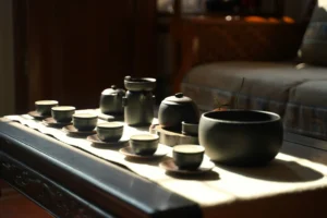A set of dark, matte ceramic teaware is elegantly arranged on a long table, bathed in warm, natural sunlight. The collection includes several small teacups on saucers, a teapot, and a large bowl, creating a serene and traditional atmosphere.
