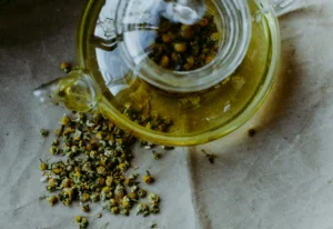 A top-down view shows a clear glass teapot filled with steeping yellow-green chamomile tea and loose blossoms in the center infuser. A small pile of dried chamomile flowers sits scattered on the crinkled brown paper background next to the teapot's spout.