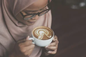A high-angle, close-up shot shows a woman wearing glasses and a light-colored hijab looking down at a white cup of coffee she is holding. The latte art on the surface of the coffee features a light bulb design surrounded by concentric half-circles.