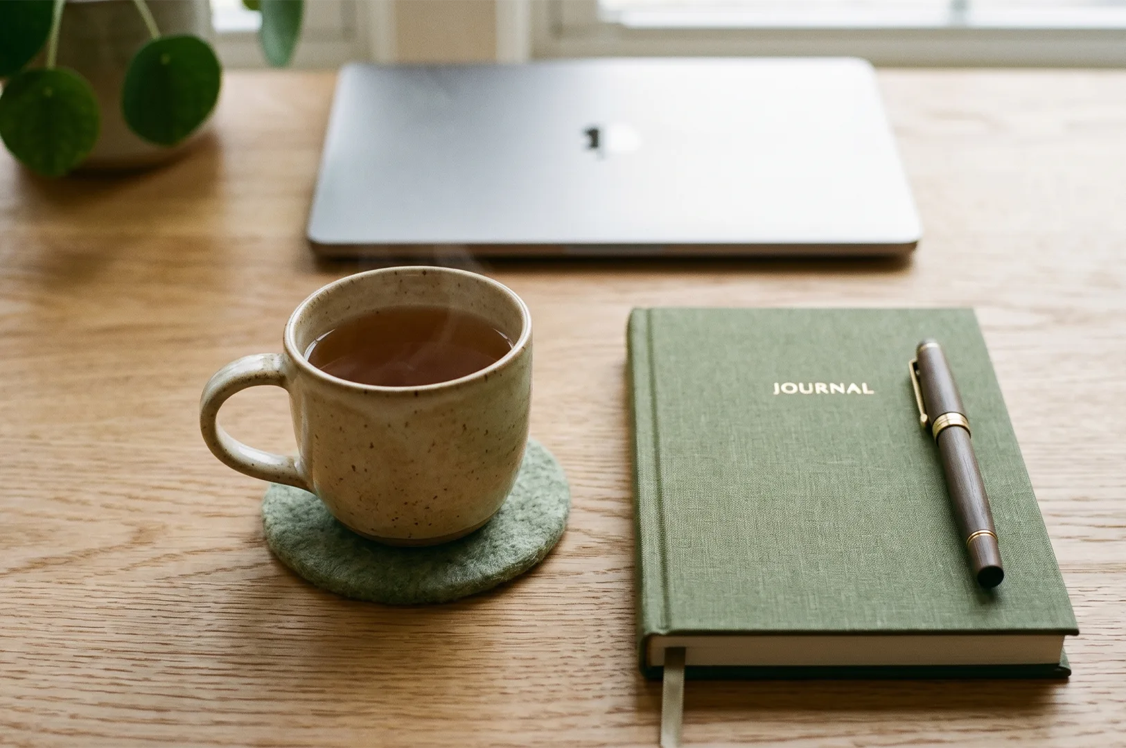 A cozy and aesthetic wooden desk setup featuring a steaming ceramic cup of herbal tea on a green felt coaster, next to a closed green fabric journal with a wooden pen, and a closed silver laptop in the background.