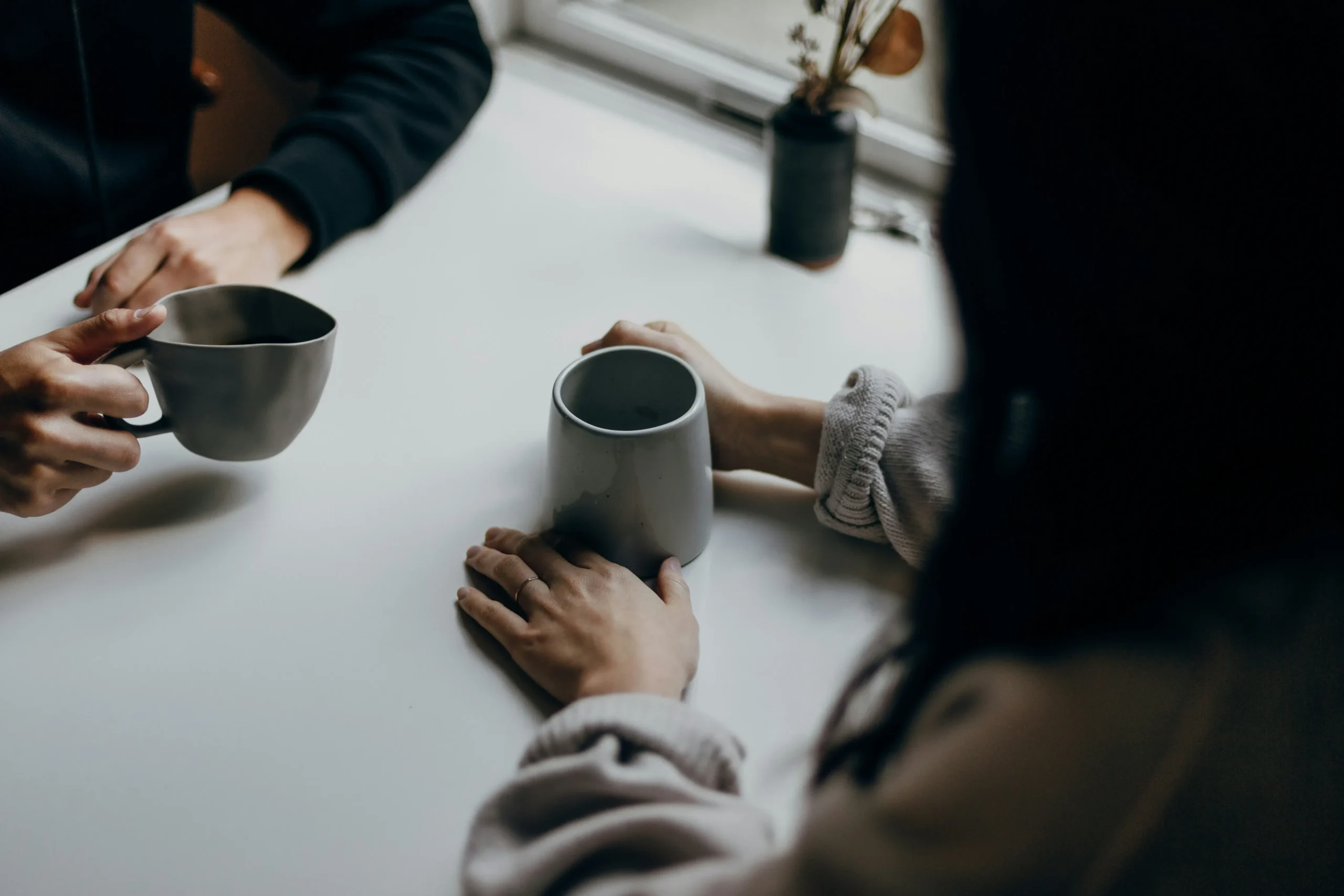 The Quiet Difference Between Drinking Tea Alone and With Someone Two people sit across from each other at a white table, their hands resting near or holding minimalist ceramic mugs. The scene is shot from a high angle with a shallow depth of field, creating a moody and intimate atmosphere.