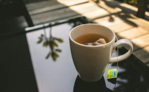 In the image, a white ceramic mug filled with tea sits on a reflective glass table outdoors. A Dilmah tea bag tag hangs from the side, while soft sunlight and shadows of foliage are visible in the background and reflections.