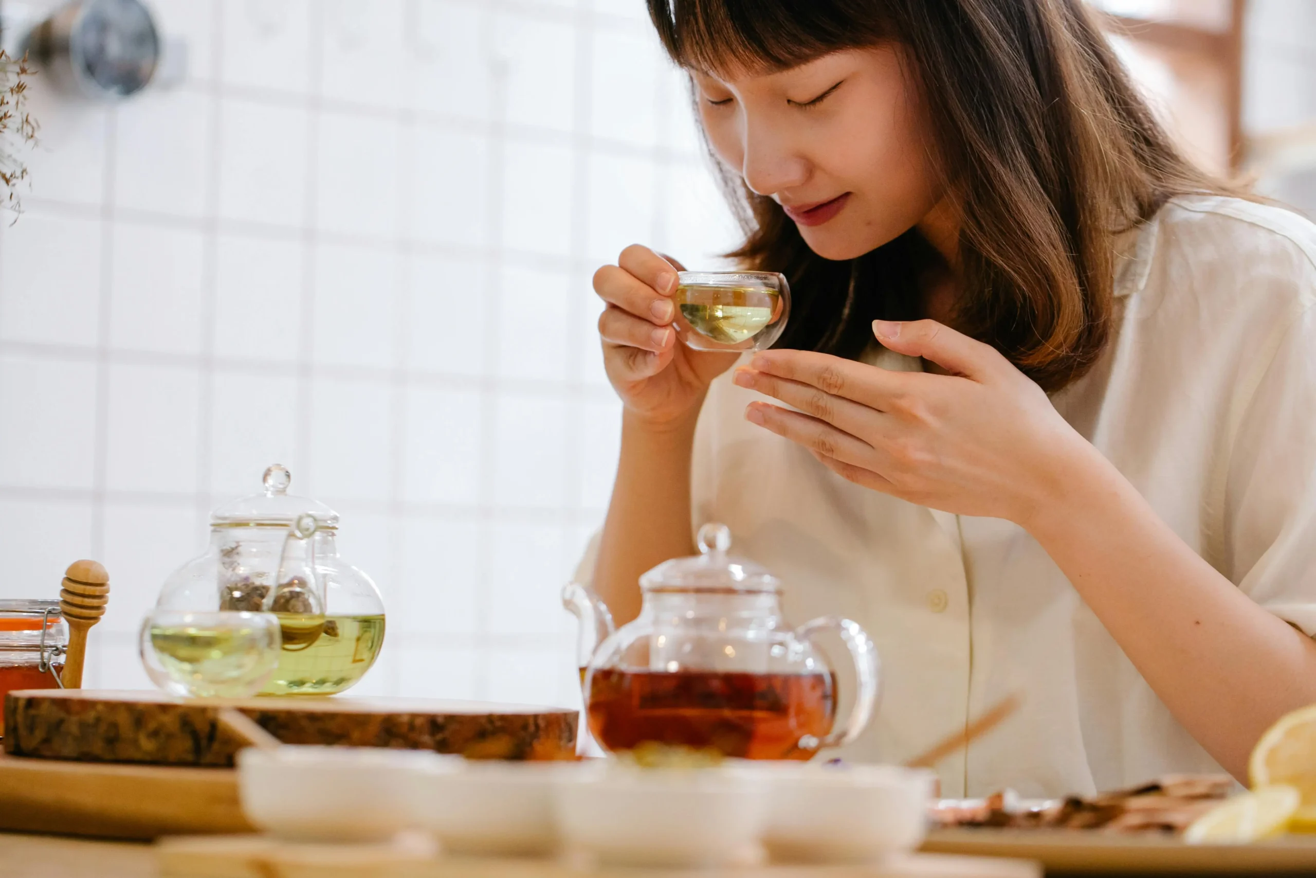The Tea We Return To Again and Again A woman is pictured holding a small glass of tea close to her face to enjoy its aroma. Several glass teapots and small bowls are arranged on the table in front of her, creating a peaceful tea-tasting scene.