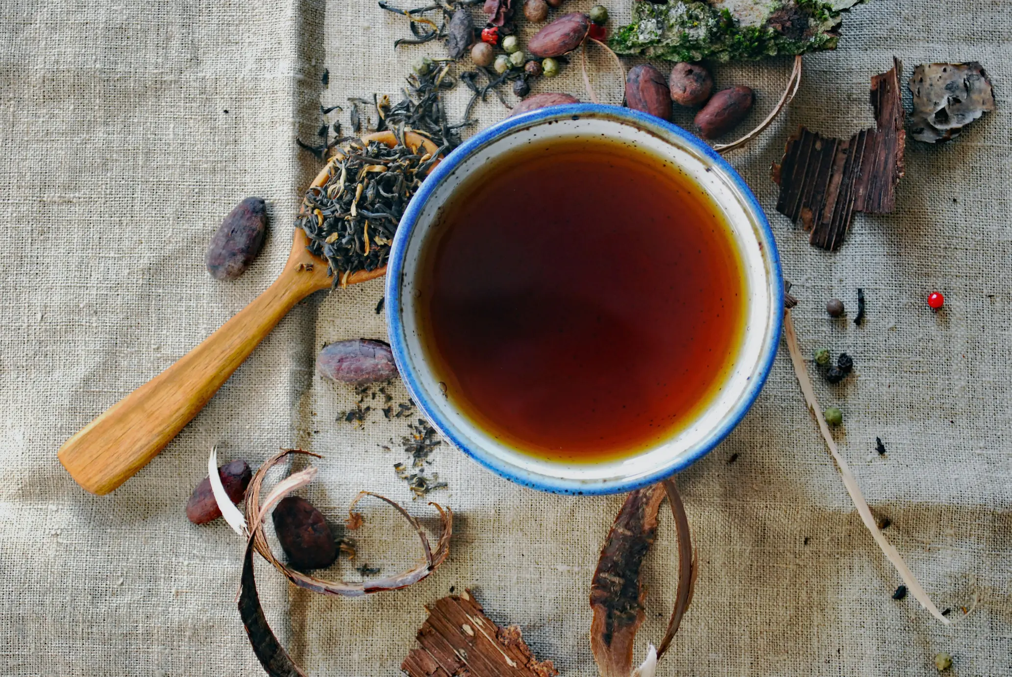 Why Oolong Tea Always Feels Slightly Mysterious An overhead shot shows a rustic ceramic bowl filled with dark amber tea, set against a textured burlap background. Surrounding the bowl are loose tea leaves on a wooden spoon, raw cacao beans, tree bark, and scattered peppercorns.